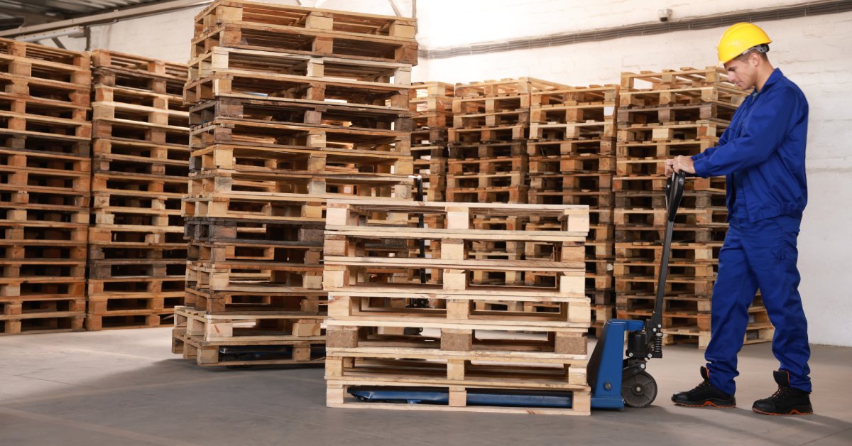 A man in a hard hat and blue jumpsuit is moving pallets with a pallet jack. Several wooden pallets are in the background.