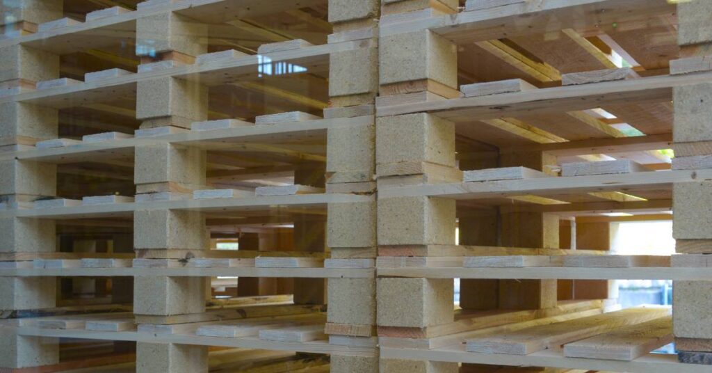 Three stacks of lightly-colored wood pallets at close range. Sunlight shines through the openings of the pallets.