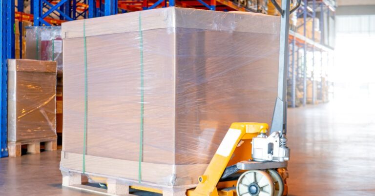 A large warehouse with orange and blue shelves. An orange pallet jack is carrying a palette with a shrink-wrapped box on top.