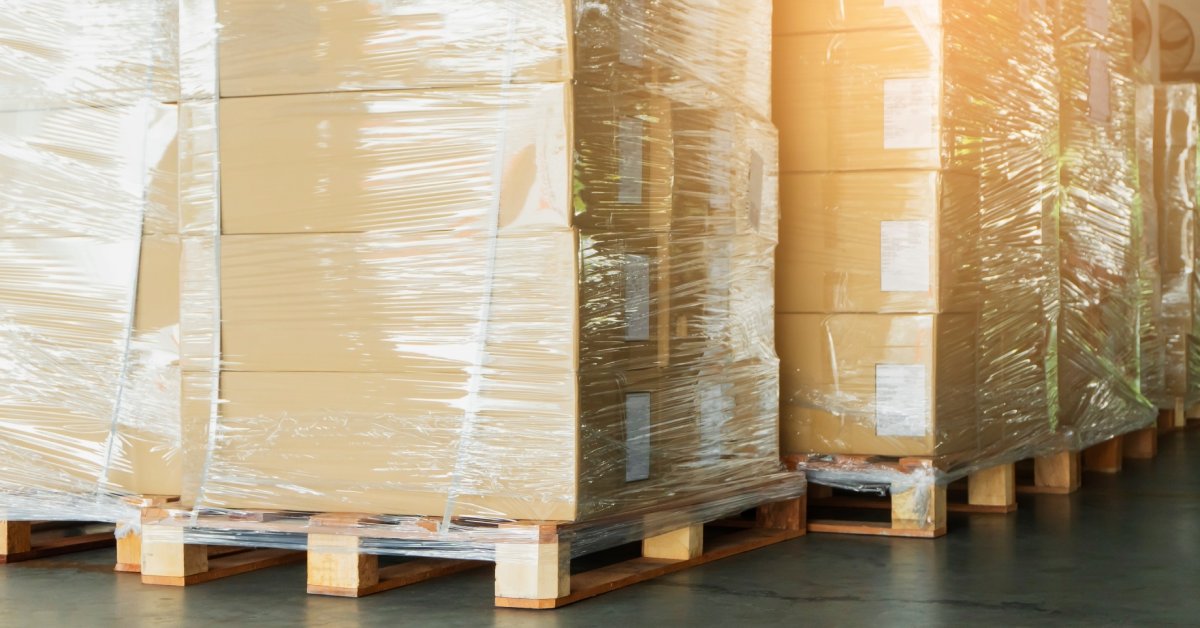 A close-up of wooden pallets on a concrete floor. All have several stacks of plastic-wrapped cardboard boxes on top of them.