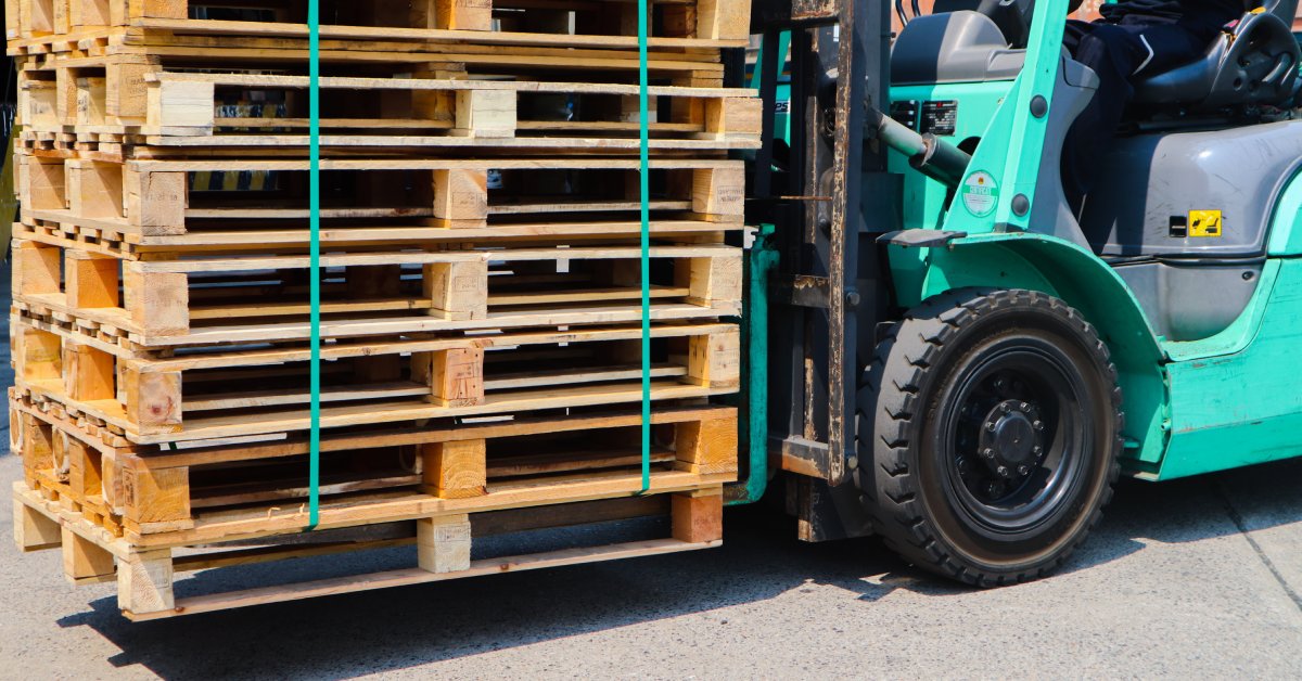 A green forklift carrying a stack of wooden pallets. The pallets are all tied together with a tight green band.