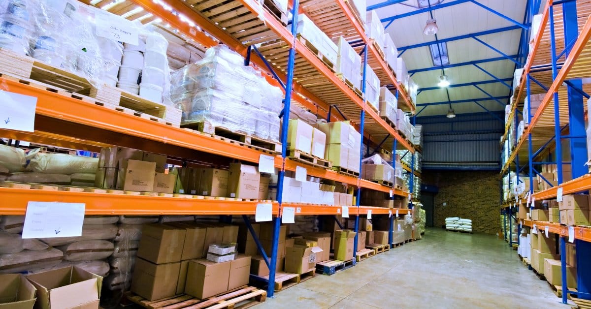 A large warehouse floor with tall orange shelves. Wooden pallets are tucked onto them with merchandise wrapped up.