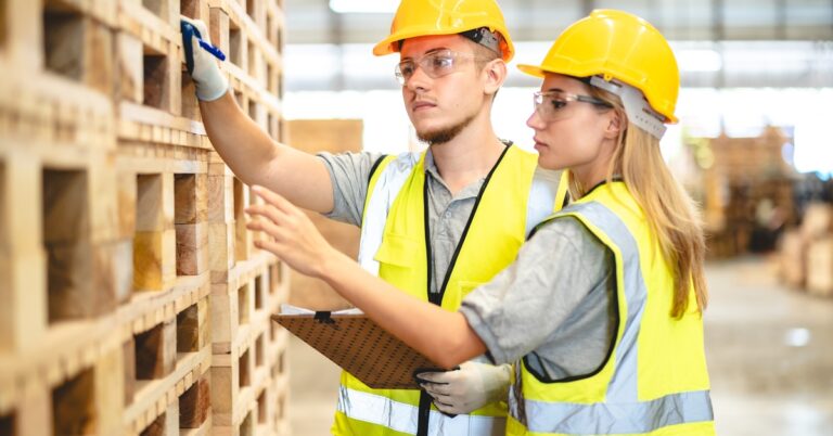 Two workers in safety vests, hardhats, and safety goggles are carefully examining a large stack of wooden pallets.
