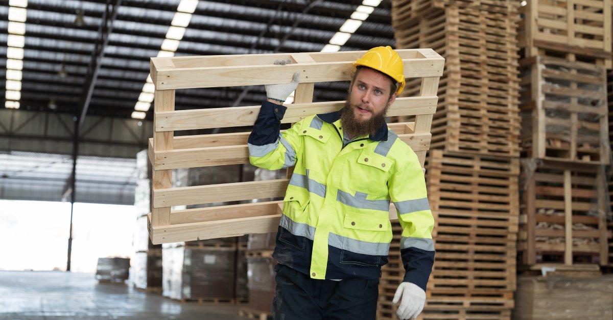 A man in a hard hat and reflective jacket is carrying a wooden pallet over his shoulder through a warehouse.