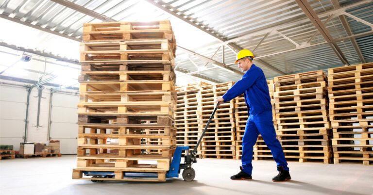 A man in a blue jumpsuit and yellow hard hat is pushing a blue pallet jack with a large stack of wooden pallets on it.