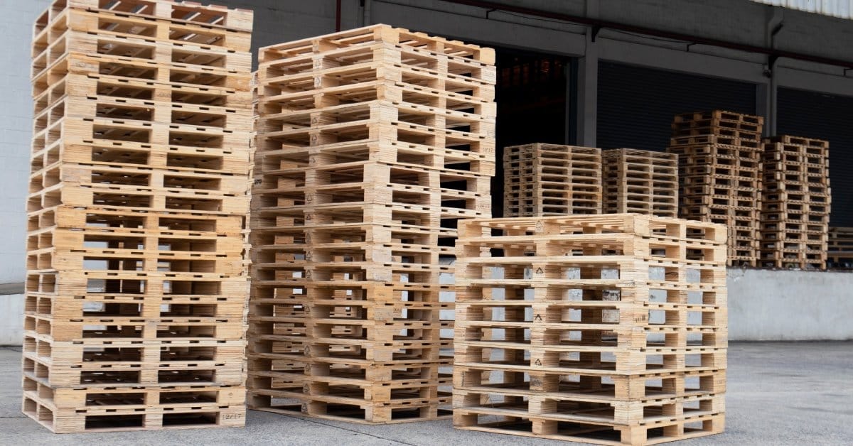 Three tall stacks of wooden pallets outside of a warehouse loading dock. More pallets are visible in the background.