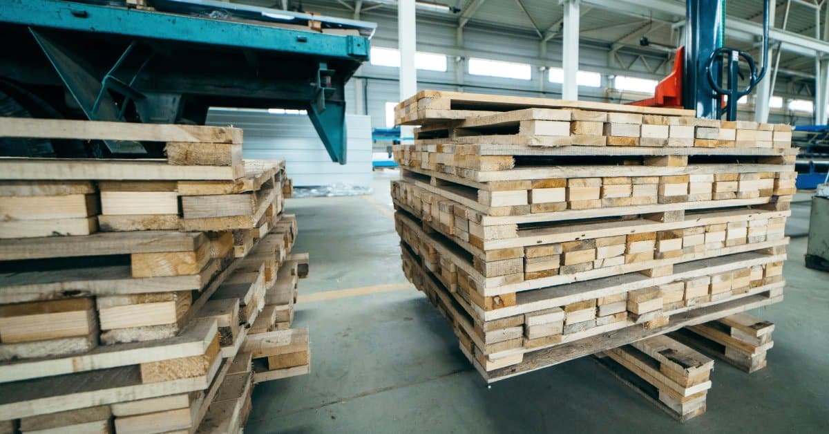 Stacks of wooden pallets resting on wooden sandwich boards to keep them off the ground. There is machinery in the background.