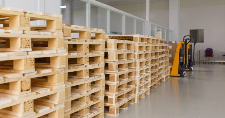 A row of pallet columns, each stacked seven to eight high, in a warehouse. Two orange pallet jacks are in the background.