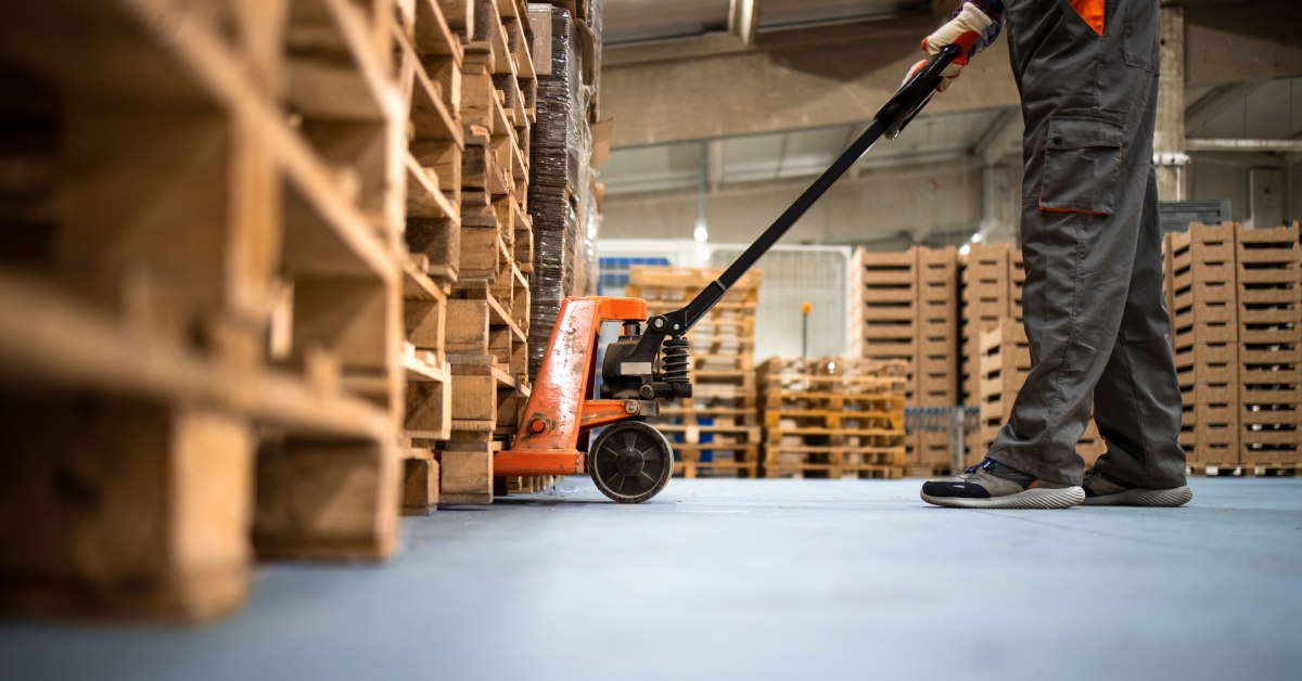 A warehouse worker in a dark gray jumpsuit. He's using an orange pallet jack to lift and move a stack of pallets.