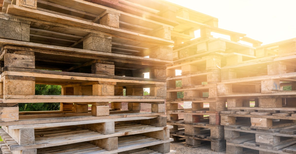 Three large stacks of wooden pallets set up outside. The sun glares on the right side. The wood is of various quality.