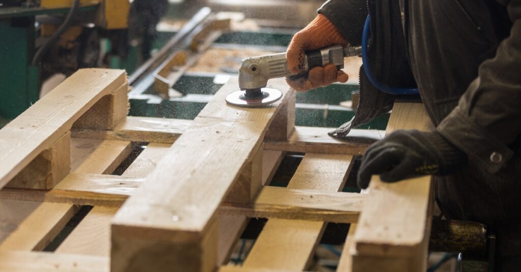 A person wearing orange and dark-green gloves uses an electronic sander to buff the surface of a wooden pallet.