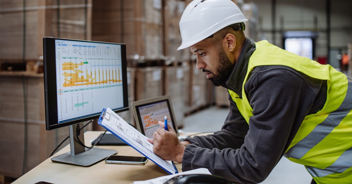 A man wearing a hard hat, neon vest, and black sweater takes notes in front of a computer with an open spreadsheet.